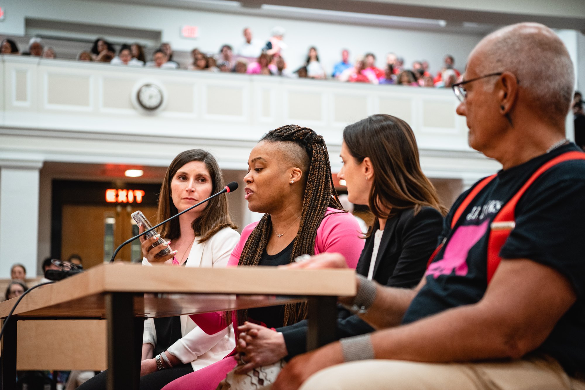 Rica Elysée speaking into a microphone at a public hearing, with a packed gallery behind her