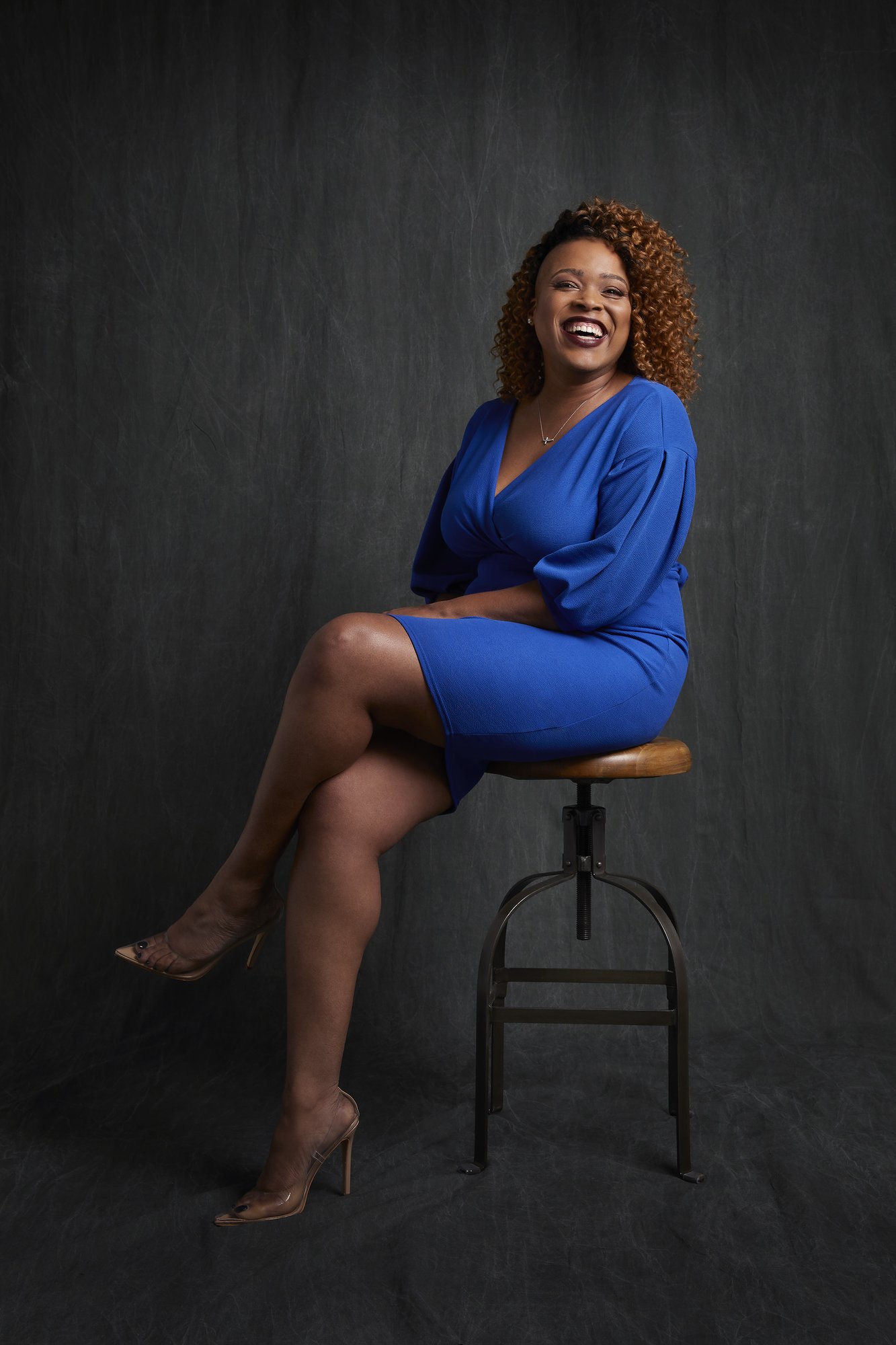 Rica Elysée smiling, seated on a stool in a blue dress, studio portrait